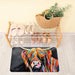 A colorful highland cow print floor mat lies on a light wood floor in front of a wooden shoe rack. The mat features a vibrant, multicolored painting of a cow's face on a dark background.